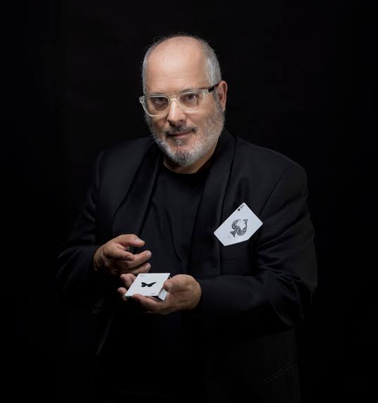 Miguel Puga, MagoMigue, vestido de negro sobre fondo oscuro, sostiene una carta con una mariposa
