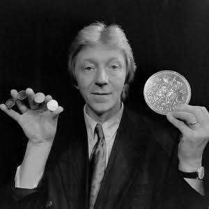 Richard Ross en blanco y negro mostrando monedas pequeñas y una moneda gigante